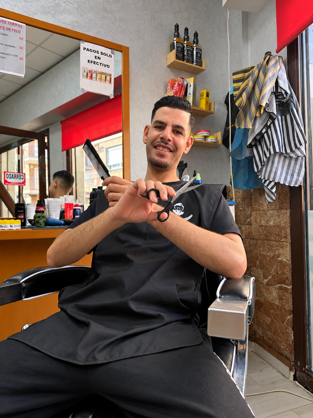 Younes sonriendo con peine y tijeras en su barbería
